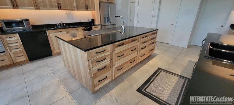 Kitchen showcasing natural hickory cabinetry with black granite countertops, a large central island with a sink, and stainless steel appliances. Project by Interstate Custom Kitchen & Bath, Inc