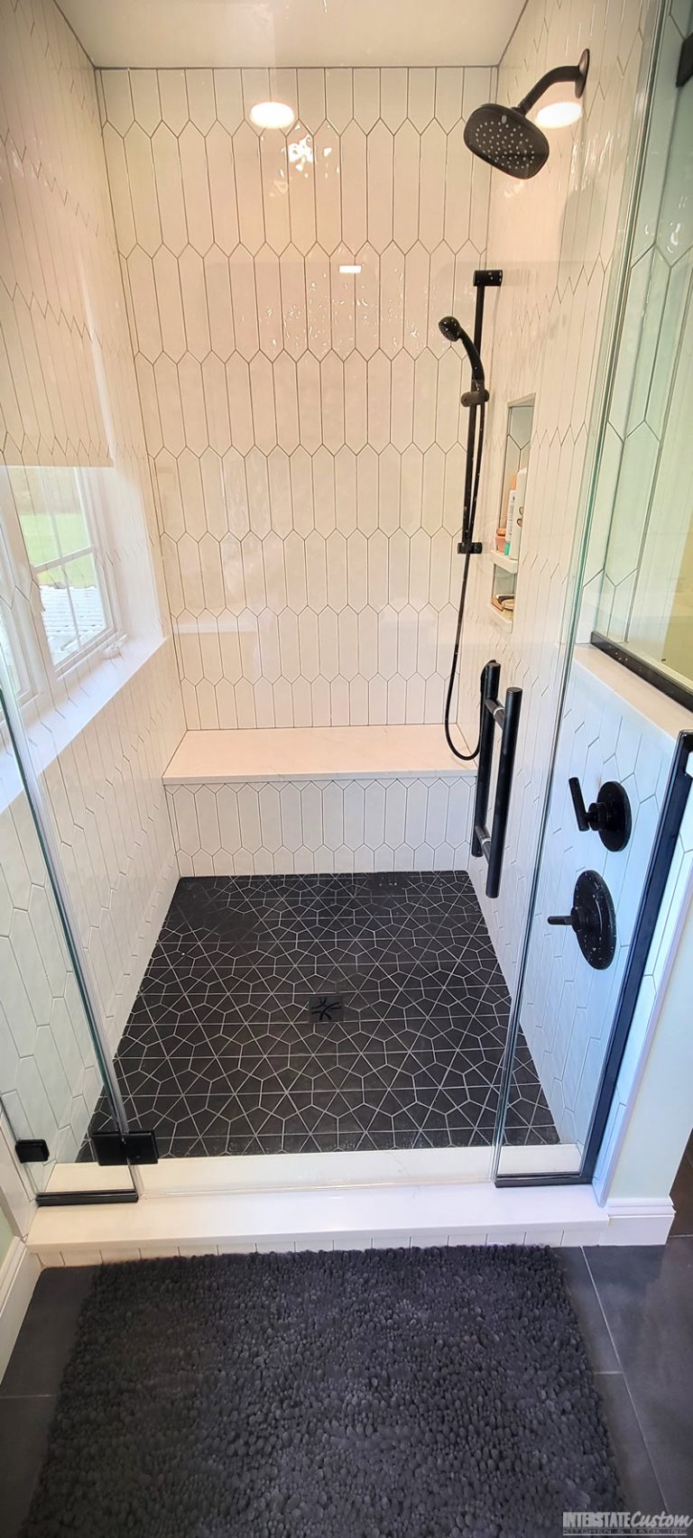 After image of a stylish shower area with white elongated hexagonal tiles, matte black fixtures including a rainfall showerhead and handheld shower, a built-in niche for toiletries, and a bench with a white quartz surface. The shower features a dark geometric patterned tile floor and glass doors with black framing, adding a modern touch.