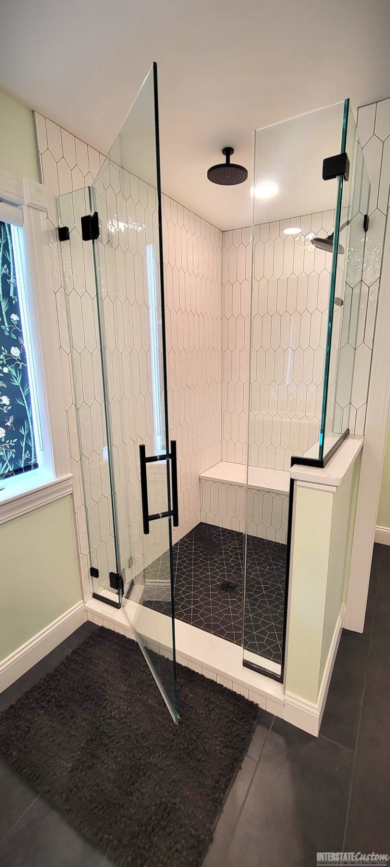 After image of a contemporary shower area featuring elongated white hexagonal tiles, matte black fixtures, a built-in bench with a white quartz surface, and a dark geometric patterned tile floor. The shower is enclosed with glass doors framed in black, creating a sleek and modern look.