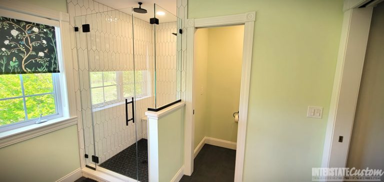 After image of a modern bathroom featuring a glass-enclosed shower with elongated white hexagonal tiles, matte black fixtures, and a dark geometric patterned tile floor. The room also includes a large window with a floral patterned shade, light green walls, and a toilet area adjacent to the shower. Project by Interstate Custom Kitchen & Bath, Inc.