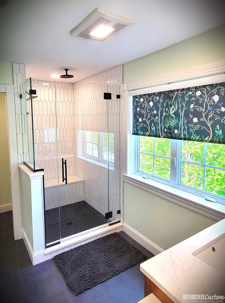 After image of a modern bathroom featuring a glass-enclosed shower with elongated white hexagonal tiles, matte black fixtures, a dark geometric patterned tile floor, and a bench with a white quartz surface. The room also includes a large window with a floral patterned shade, light green walls, and dark tile flooring. Project by Interstate Custom Kitchen & Bath, Inc.