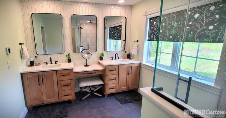 After image of a modern bathroom featuring a double vanity with wood cabinetry and white quartz countertops, black fixtures, elongated white hexagonal tile backsplash, and three large mirrors with black frames. The vanity includes a makeup station with a cushioned stool, and the room is illuminated by natural light from a large window with a floral patterned shade. Project by Interstate Custom Kitchen & Bath, Inc.