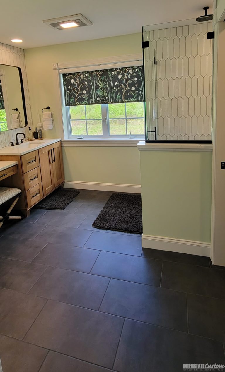 After image of a modern bathroom featuring a double vanity with wood cabinetry and white quartz countertops, black fixtures, elongated white hexagonal tile backsplash, and a large window with a floral patterned shade. The room also includes a spacious glass-enclosed shower and dark tile flooring. Project by Interstate Custom Kitchen & Bath, Inc.