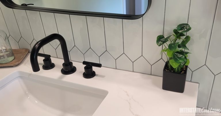 Close-up of a modern bathroom vanity featuring a sleek white countertop with subtle veining, matte black fixtures, a hexagonal tile backsplash in light gray, a rectangular mirror with a black frame, and a small green potted plant. Project by Interstate Custom Kitchen & Bath, Inc.