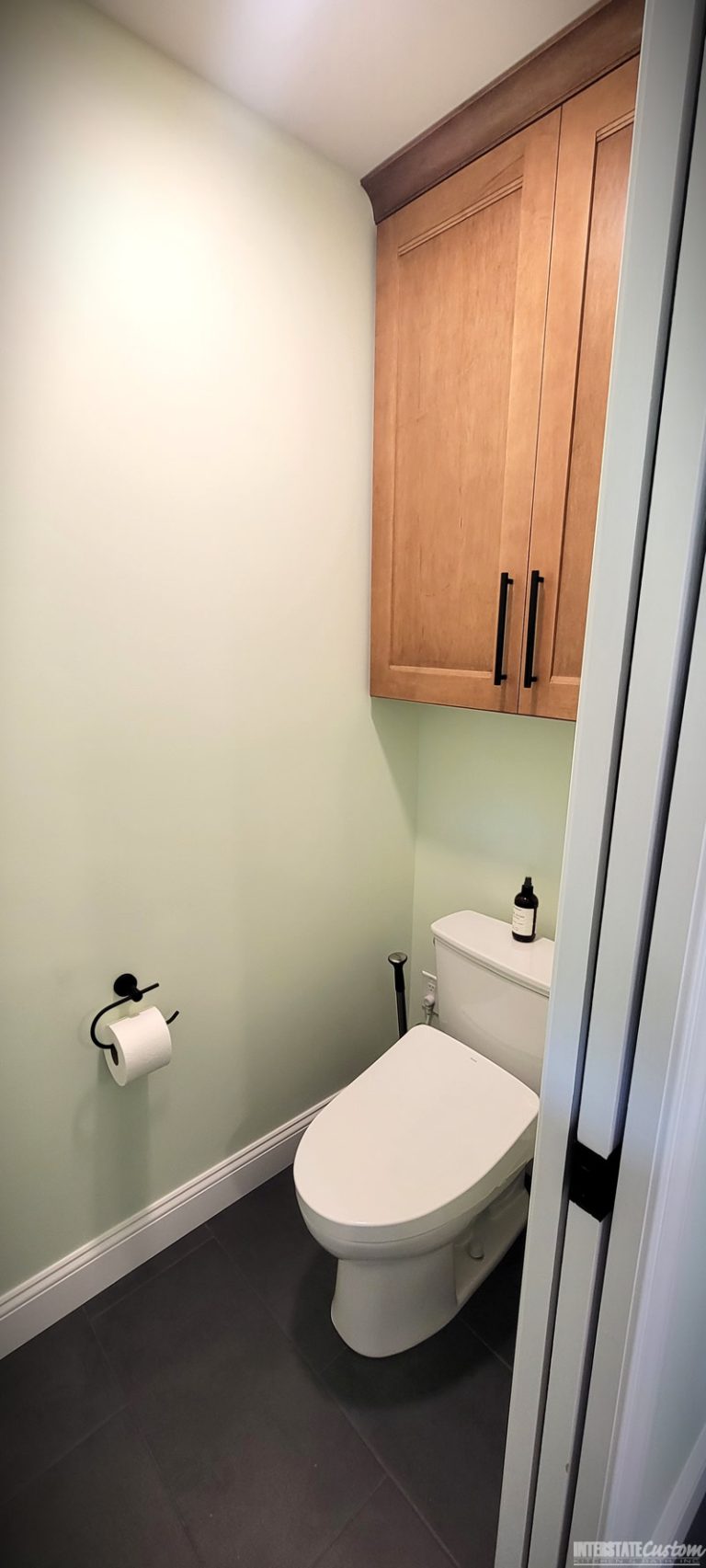 After image of a modern bathroom toilet area featuring a sleek white toilet, light green walls, dark tile flooring, and wood cabinetry with black handles for additional storage. The minimalistic design creates a clean and contemporary look. Project by Interstate Custom Kitchen & Bath, Inc.