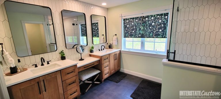 After image of a modern bathroom featuring a double vanity with wood cabinetry and white quartz countertops, black fixtures, elongated white hexagonal tile backsplash, and three large mirrors with black frames. The vanity includes a makeup station with a cushioned stool, and the window has a floral patterned shade. The dark tile floor complements the overall contemporary design. Project by Interstate Custom Kitchen & Bath, Inc.