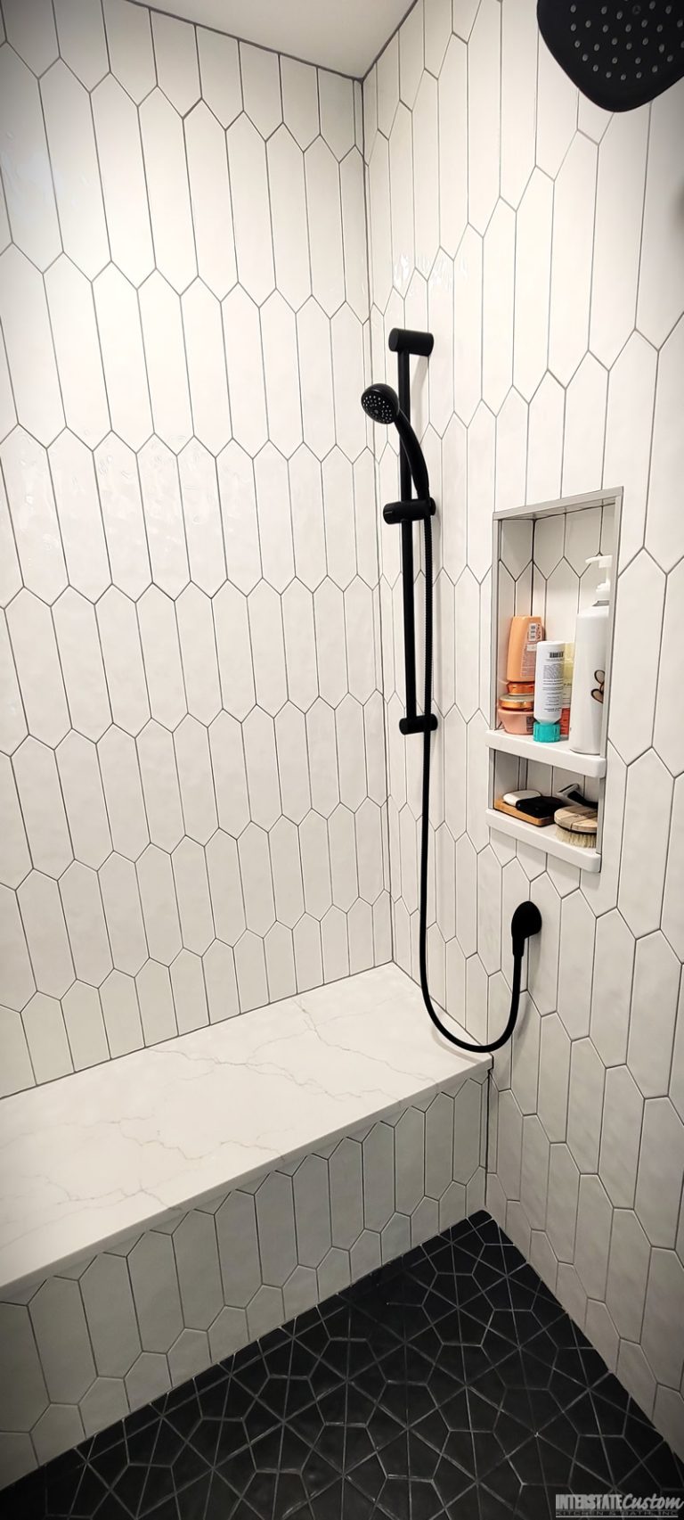 After image of a modern shower area with elongated white hexagonal tiles, matte black fixtures including a rainfall showerhead and handheld shower, a built-in niche for toiletries, a bench with a white quartz surface, and a dark geometric patterned tile floor. The shower is enclosed with glass doors framed in black