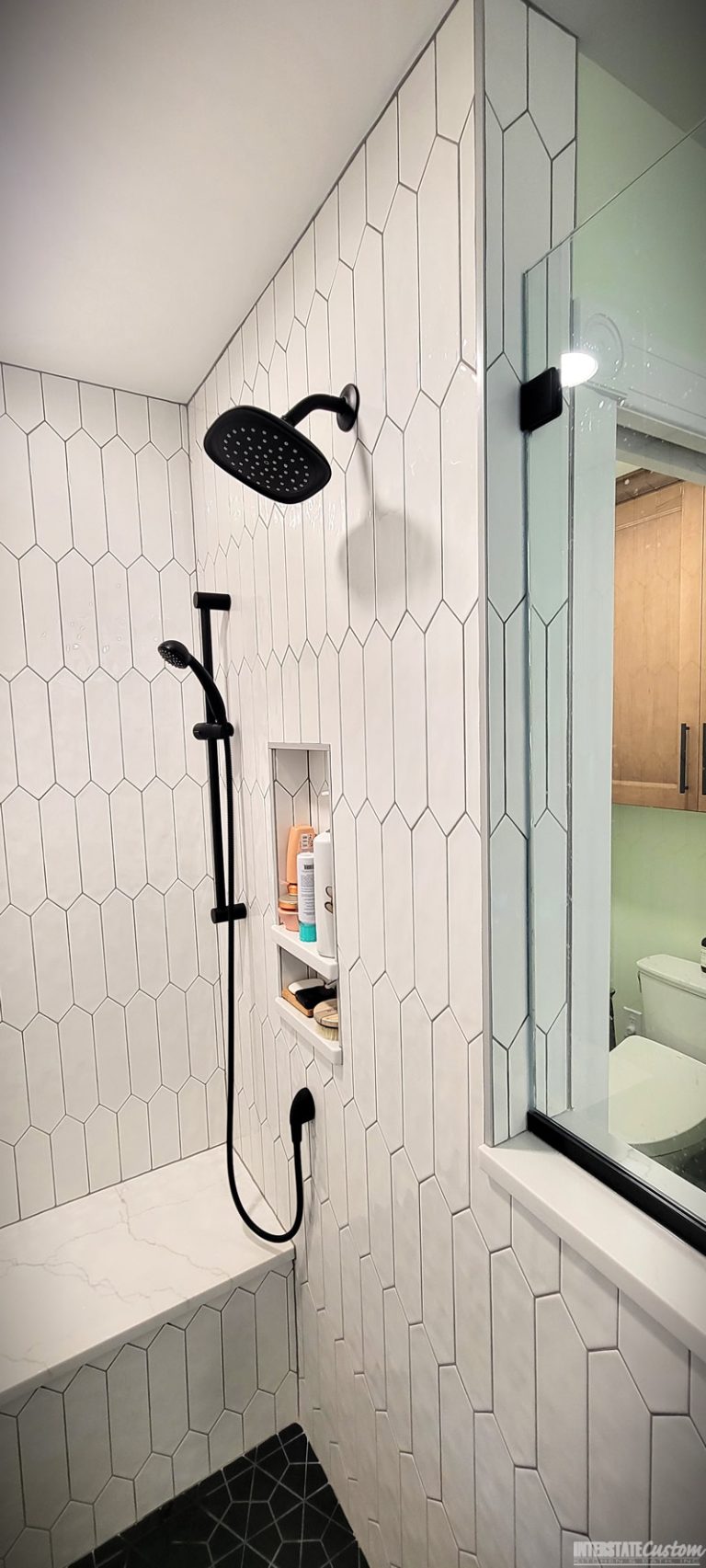 modern shower area featuring white elongated hexagonal tiles, matte black fixtures including a rainfall showerhead and a handheld shower, a built-in niche with shelves for toiletries, and a bench with a white quartz surface. The shower area is adjacent to a glass partition with black trim, providing a sleek and contemporary look. Project by Interstate Custom Kitchen & Bath, Inc.