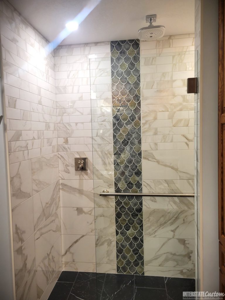 Shower area in a bathroom remodel featuring marble-look wall tiles with subtle gray veining, an eye-catching vertical strip of iridescent fish scale tiles in green and blue hues, a modern square rainfall showerhead, a polished chrome grab bar, and a glass partition. Project by Interstate Custom Kitchen & Bath, Inc.