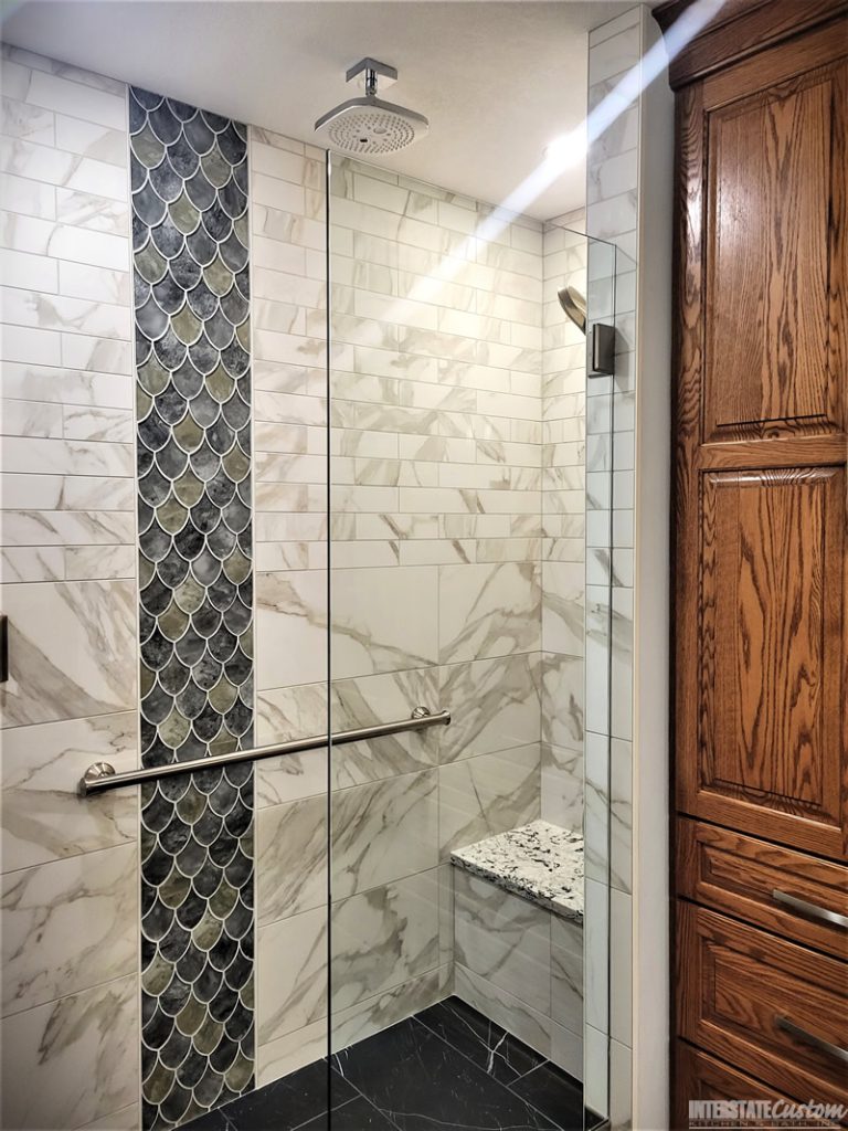 Shower area in a bathroom remodel featuring marble-look wall tiles with subtle gray veining, an eye-catching vertical strip of iridescent fish scale tiles in green and blue hues, a modern square rainfall showerhead, a polished chrome grab bar, and a built-in bench with a matching quartz surface. Project by Interstate Custom Kitchen & Bath, Inc.