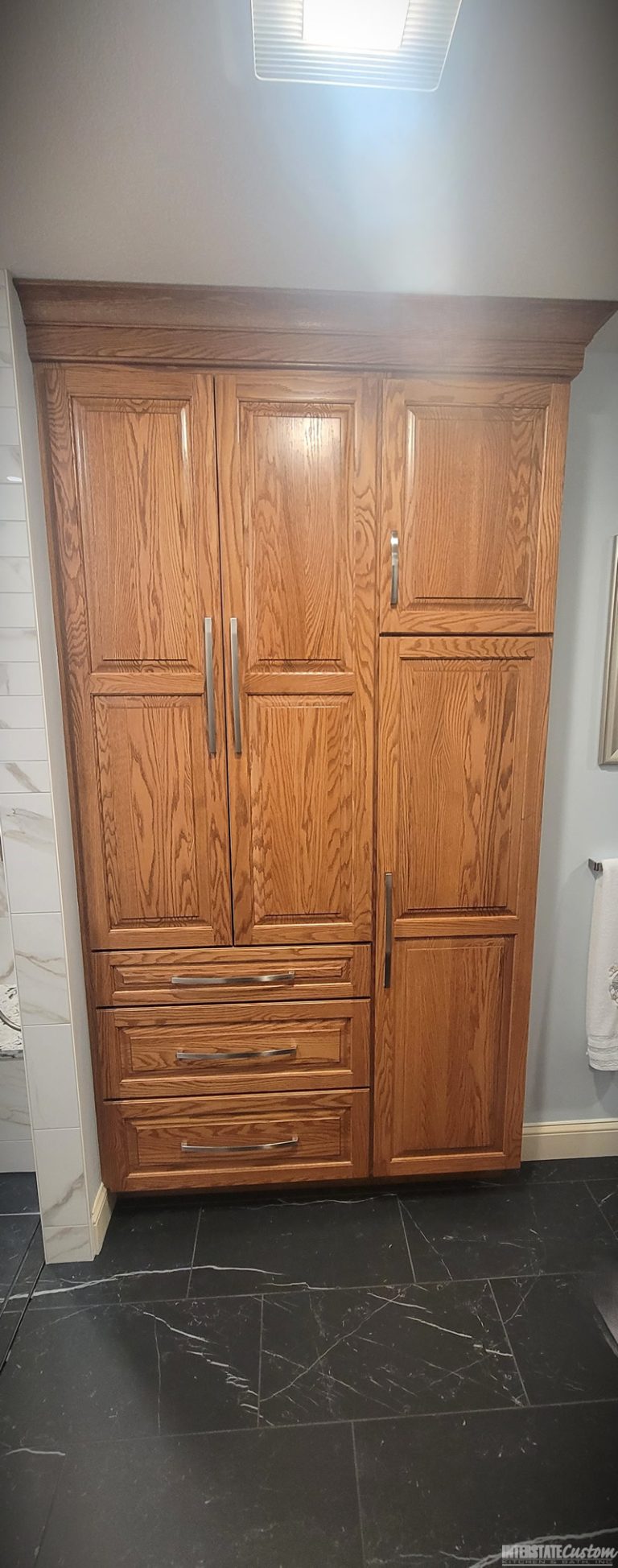 Custom oak cabinetry in a bathroom remodel featuring ample storage with multiple cabinets and drawers, sleek silver handles, and rich, natural wood grain, complemented by dark tile flooring with subtle white veining. Project by Interstate Custom Kitchen & Bath, Inc.