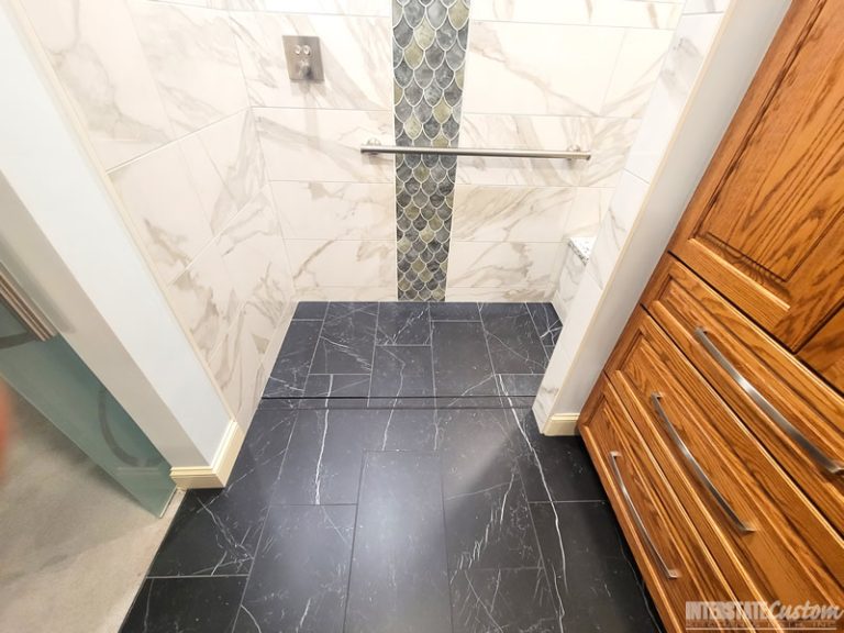 View of the shower floor in a bathroom remodel featuring dark, large-format tiles with subtle white veining, complementing the marble-look wall tiles. A vertical strip of iridescent fish scale tiles in green and blue hues adds a striking accent. The shower includes a polished chrome grab bar and a built-in bench with a matching quartz surface. Project by Interstate Custom Kitchen & Bath, Inc.