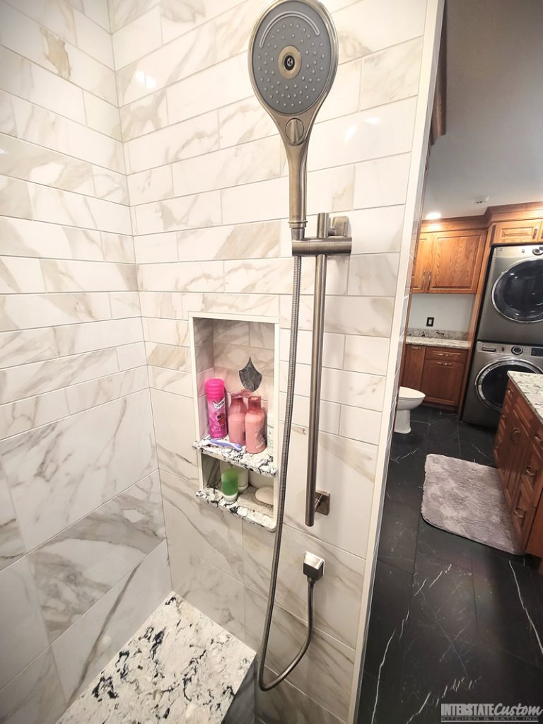 Close-up of a shower area in a bathroom remodel featuring large-format marble-look tiles with gray veining, a modern handheld shower fixture in polished chrome, a built-in niche with shelves showcasing a mix of practical storage and decorative fish scale tile accents, and a bench with a matching quartz surface. Project by Interstate Custom Kitchen & Bath, Inc.