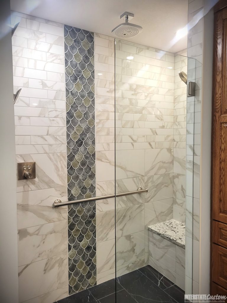 Elegant shower area in a bathroom remodel featuring large-format marble-look tiles with subtle gray veining, a striking vertical strip of iridescent fish scale tiles in deep greens and blues, a square rainfall showerhead, a sleek handheld shower, a glass partition, a polished chrome grab bar, and a built-in bench with a matching quartz surface. Project by Interstate Custom Kitchen & Bath, Inc.