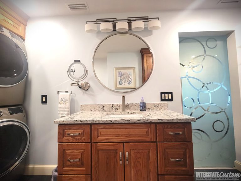 Vanity area in a bathroom remodel featuring oak cabinetry with ample storage, a luxurious quartz countertop with white and gray veining, a round frameless mirror, contemporary light fixtures, a convenient magnifying mirror, and a decorative glass partition with a circular pattern. Project by Interstate Custom Kitchen & Bath, Inc.