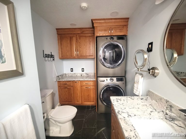 Bathroom remodel featuring a compact laundry area with a stacked washer and dryer, warm oak cabinetry, luxurious quartz countertops with white and gray veining, a convenient magnifying mirror, and dark tile flooring, completed by Interstate Custom Kitchen & Bath, Inc.