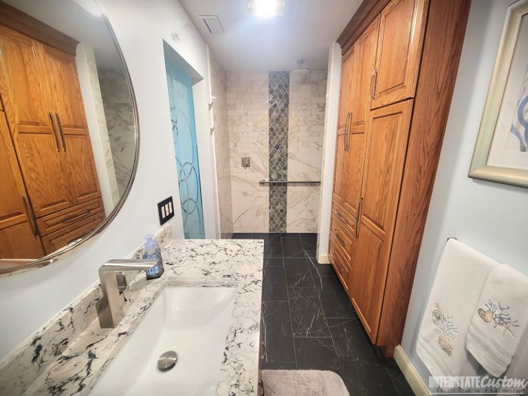 Bathroom remodel featuring a seamless integration of a vanity, shower, and custom cabinetry. The vanity includes a luxurious quartz countertop with white and gray veining, modern chrome fixtures, and a round frameless mirror. The shower area is highlighted by marble-look wall tiles with a vertical strip of iridescent fish scale tiles in green and blue hues, a square rainfall showerhead, and a polished chrome grab bar. Custom oak cabinetry provides ample storage, and the dark tile flooring with subtle white veining unifies the space. Project by Interstate Custom Kitchen & Bath, Inc.