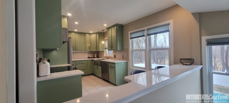 Wide-angle view of a completed kitchen refacing project with green cabinetry, stainless steel appliances, white countertops, and a tiled backsplash, featuring large windows and a breakfast bar. Project by Interstate Custom Kitchen & Bath, Inc.