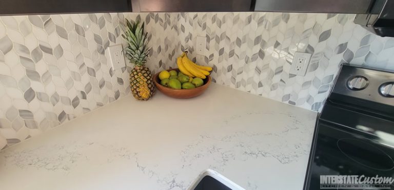 Detailed view of the natural marble braided mosaic tile backsplash and Hanstone Chantilly lace quartz countertops with a fruit bowl in the Simply Elegant kitchen remodel. Project by Interstate Custom Kitchen & Bath, Inc.