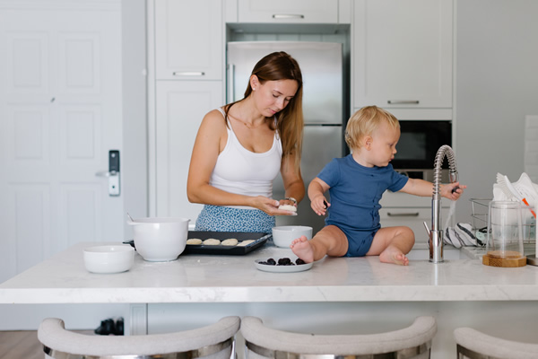 Mother and toddler in a modern kitchen, with the mother preparing food and the toddler sitting on a white porcelain countertop next to a sink by Interstate Custom Kitchen & Bath, Inc